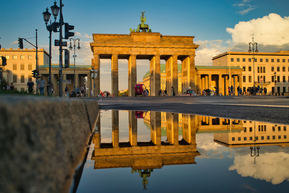 Das Brandenburger Tor in Berlin in der goldenen Abendsonne, aufgenommen aus Richtung vom Platz des 18. März. Im Vordergrund spiegelt sich das Bauwerk samt Quadriga im tiefstehenden Licht klar in einer Pfütze auf der Straße.